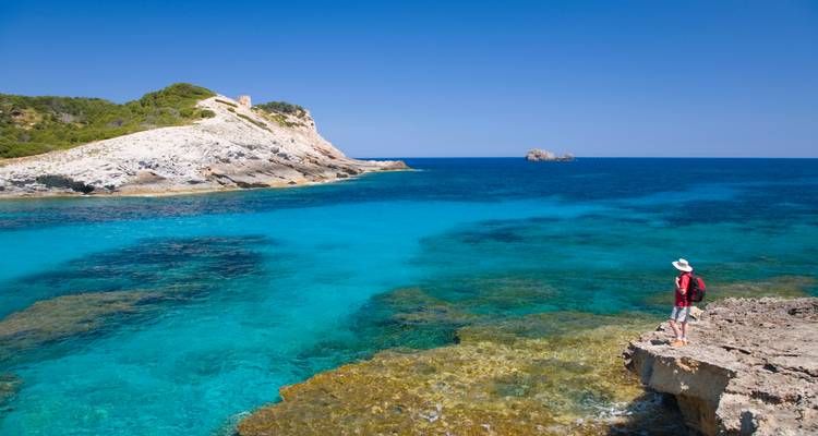 Ein einsamer Wanderer mit Rucksack steht auf einem felsigen Vorsprung und blickt auf leuchtend türkisfarbenes Mittelmeer und ein kleines weißes Felsenvorgebirge unter klarem blauem Himmel.