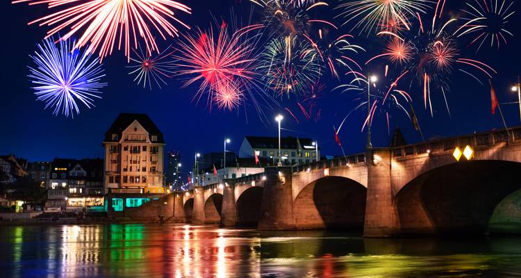 Buntes Feuerwerk explodiert über einer alten Steinbrücke, die sich in einem Fluss an einem festlichen Abend spiegelt.