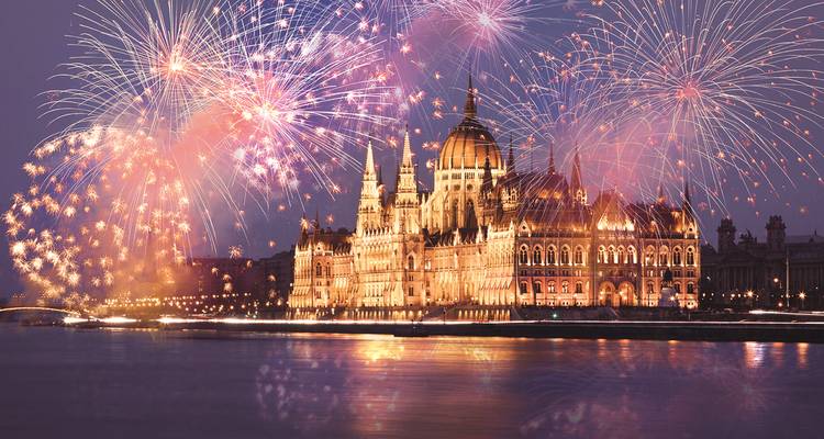 Bâtiment du Parlement hongrois illuminé se reflétant dans le Danube sous un ciel rempli de feux d'artifice colorés.