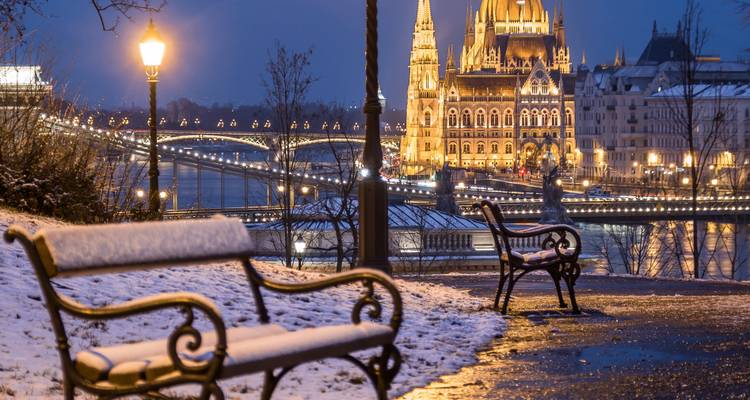Scène nocturne hivernale du Parlement de Budapest illuminé au bord du Danube, vue depuis un sentier enneigé en bordure de fleuve.