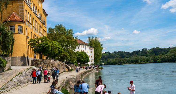 Fußgänger flanieren entlang einer Uferpromenade neben bunten historischen Gebäuden und der breiten Donau an einem strahlend sonnigen Tag.