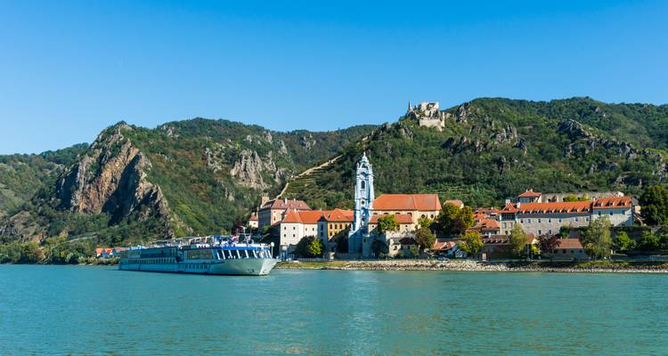 Blau bekrönter Kirchturm und rotgedecktes Dorf Dürnstein, eingerahmt von steilen grünen Hügeln und einem vorbeifahrenden Kreuzfahrtschiff auf der Donau.