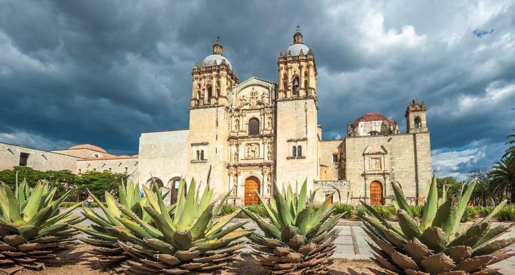 Église Santo Domingo à Oaxaca encadrée par des plants d'agave sous des nuages dramatiques.