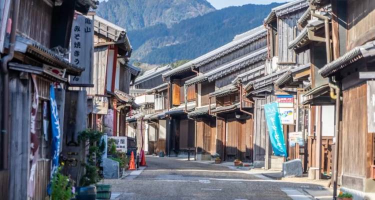 Ruelle paisible bordée de bâtiments en bois bien préservés de la période Edo avec des montagnes lointaines sous un ciel dégagé.