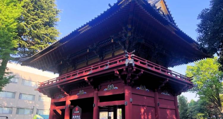 Tour de porte bouddhiste peinte en rouge traditionnel entourée d'arbres feuillus sous un ciel bleu dégagé.
