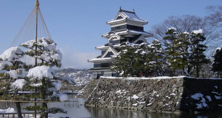 Le château de Matsumoto aux pics enneigés se reflétant dans une douve tranquille avec des arbres hivernaux.