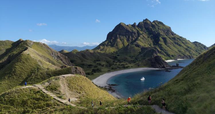 Les crêtes vertes de l'île de Padar descendent vers une plage en croissant et un yacht amarré sous un ciel dégagé.