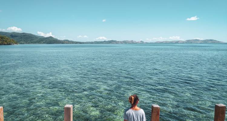 Un voyageur est assis sur une jetée en bois contemplant une mer turquoise cristalline sous un ciel lumineux.