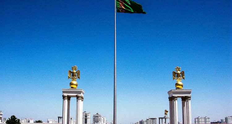 Drapeau turkmène flottant au-dessus de grandes colonnes blanches surmontées d'aigles dorés contre un ciel bleu clair.