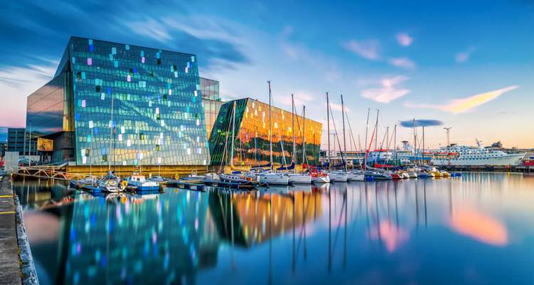 La salle de concert en verre Harpa de Reykjavik reflète les cieux pastel du soir à côté d'une marina paisible.