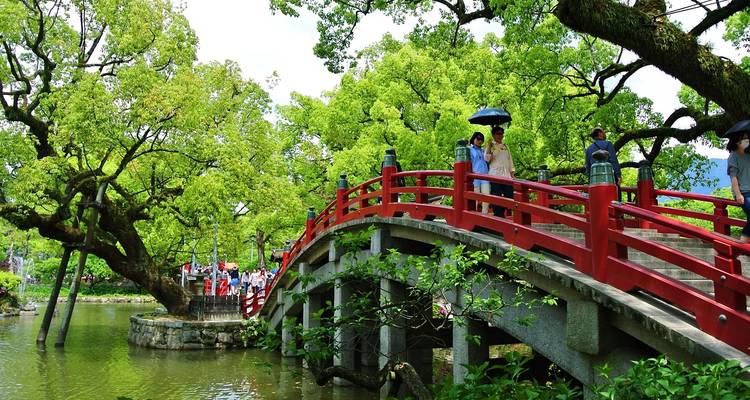 Un puente de madera rojo tradicional con arcos que cruza un estanque rodeado de árboles verdes vibrantes, con visitantes llevando paraguas.