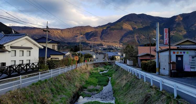 Camino de un tranquilo pueblo rural japonés que sigue un arroyo estrecho con montañas de fondo bajo la luz del atardecer.