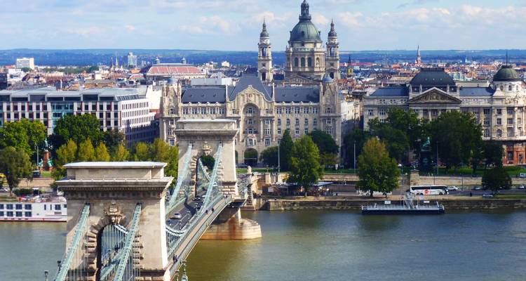 El Puente de las Cadenas de Budapest cruza el Danubio con la Basílica de San Esteban y el horizonte de la ciudad bajo una luz diurna brillante.