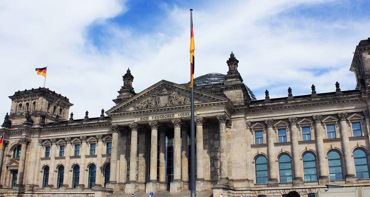 La majestuosa fachada del edificio Reichstag de Berlín con banderas alemanas ondeando bajo un cielo azul y blanco dramático.
