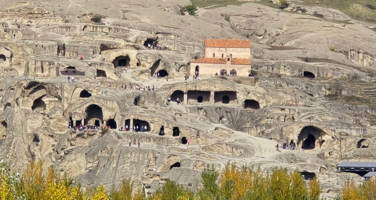 Ancient cave city carved into a rocky hillside with small church and visitors exploring the terraces.