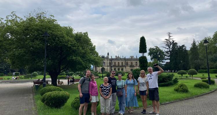 Smiling tour group posing in front of lush gardens and historic palace on an overcast day.