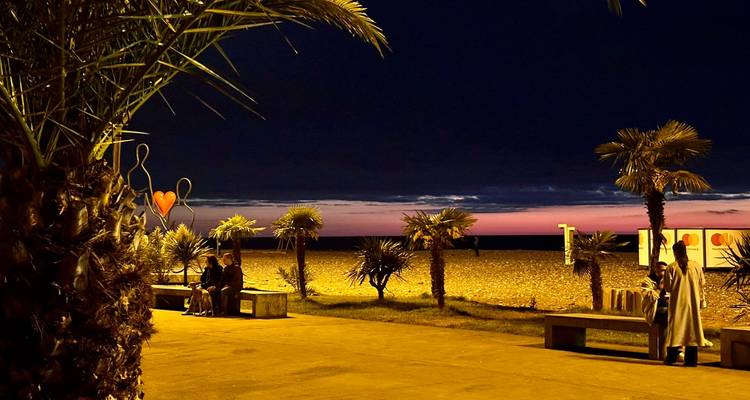 Nighttime beach promenade with silhouetted palm trees and small groups relaxing by the sea.