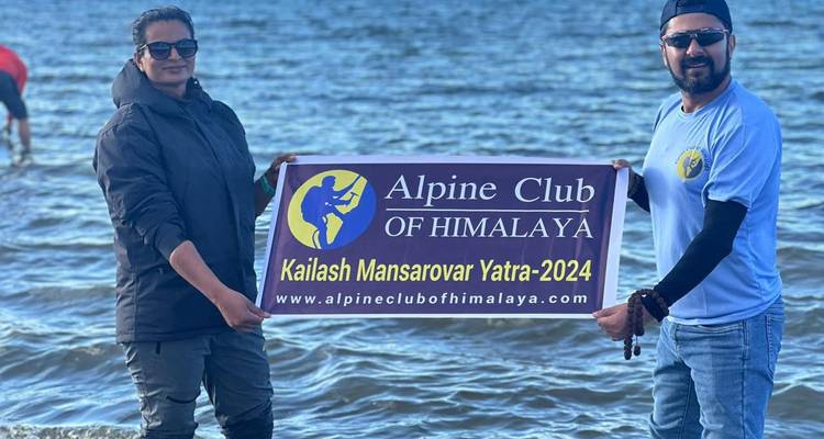 Two travellers standing in shallow lake water holding a banner for Kailash Mansarovar Yatra 2024