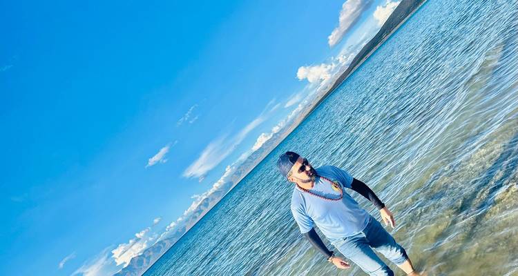 Man wading through crystal-clear blue lake under a vibrant sky with distant mountains