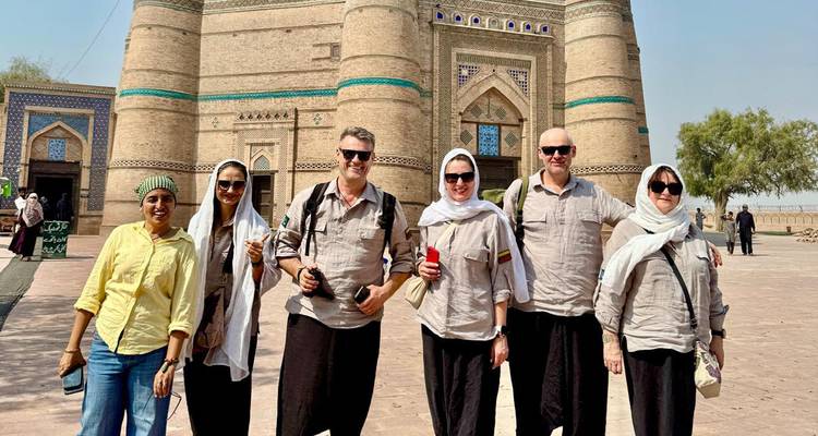 Des voyageurs se tiennent devant un mausolée orné en brique avec des carreaux turquoise sous le soleil éclatant du désert.