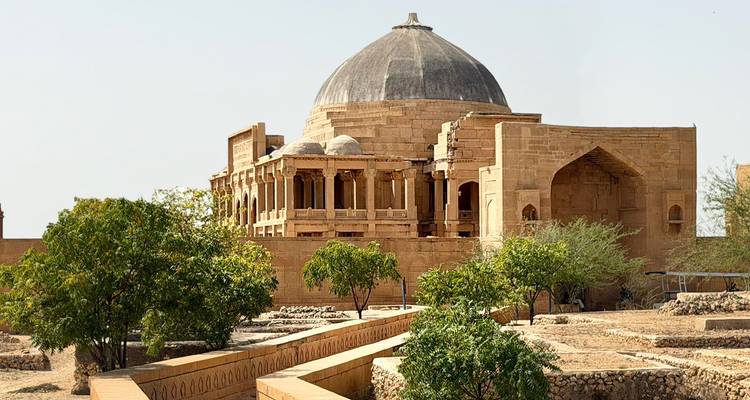 Mausolée isolé en grès avec toit en dôme entouré de terrasses paysagées arides et d'arbres clairsemés.