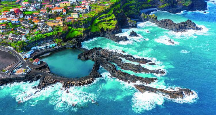 Vue aérienne spectaculaire des piscines naturelles de lave de Porto Moniz entourées de vagues turquoise qui se brisent et d'un village perché sur la falaise.