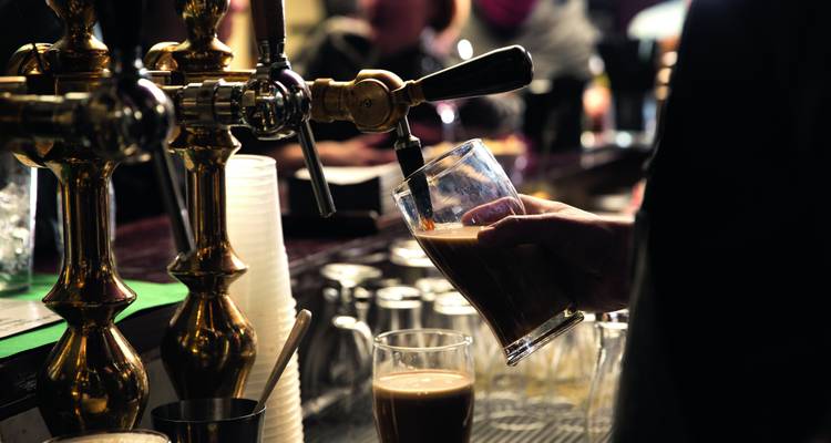 Barkeeper schenkt dunkles Stout aus Messinghähnen in ein Pintglas in einer lebhaften irischen Kneipe