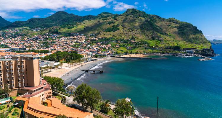 A panoramic view of a green mountain backdrop, a sweeping bay and a seaside town with black-sand beach.