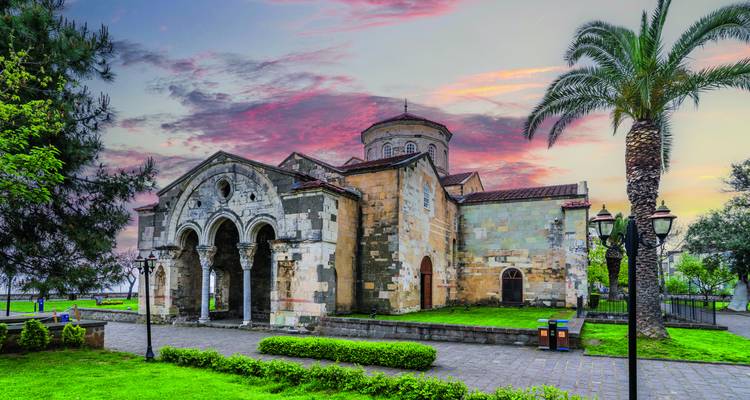 Steinkirche in Trabzon, umgeben von Grün mit einer Palme und farbenfrohen, von der Abendsonne beleuchteten Wolken darüber.