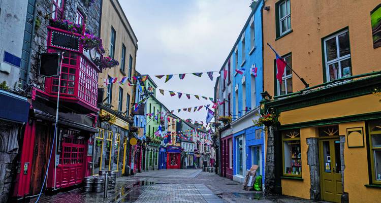 Bunte schmale Straße gesäumt von bemalten Geschäften, Wimpelfahnen und überfließenden Blumenkästen nach einem leichten Regen.