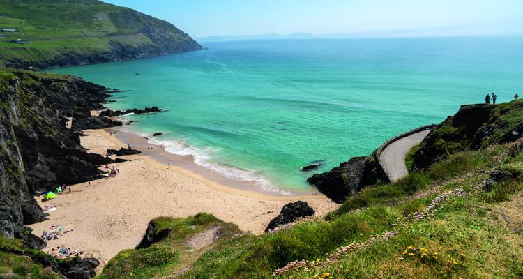 Türkise Bucht mit Sandstrand, gewundener Küstenstraße und sanften grünen Landzungen unter klarem Himmel.