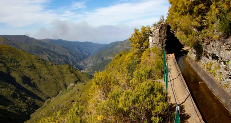 Schmale Levada-Bewässerungskanäle säumen einen Berghang mit Blick auf tiefgrüne Täler unter flächig verteilten Wolken