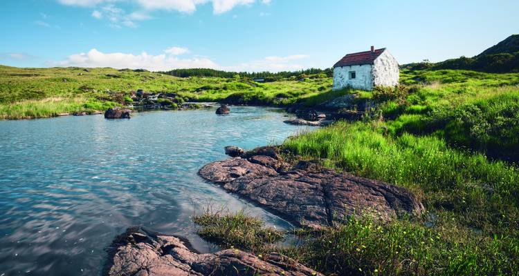 Abgelegenes weiß getünchtes Häuschen neben einem ruhigen Teich und üppigem grünem Moorland unter klarem blauem Himmel.