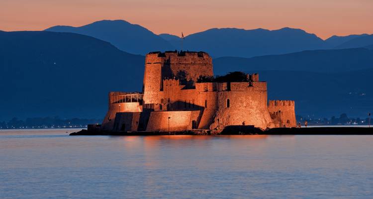Forteresse maritime de Bourtzi illuminée dans des teintes chaudes de crépuscule sur un fond de montagnes.