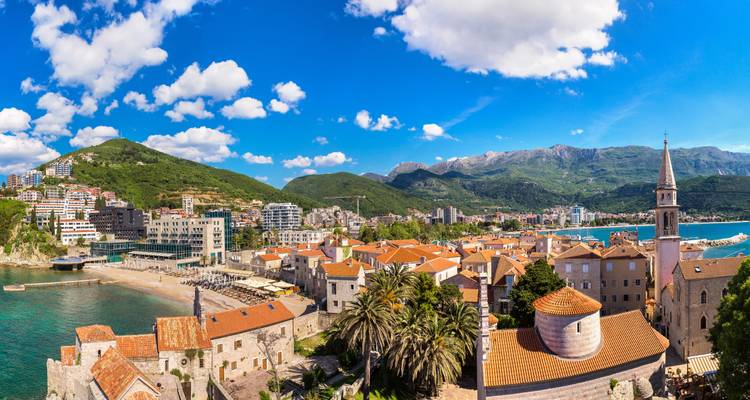 Panoramablick über die Terrakotta-Dächer der Altstadt von Budva, Palmen und die türkisfarbene Adria-Bucht.