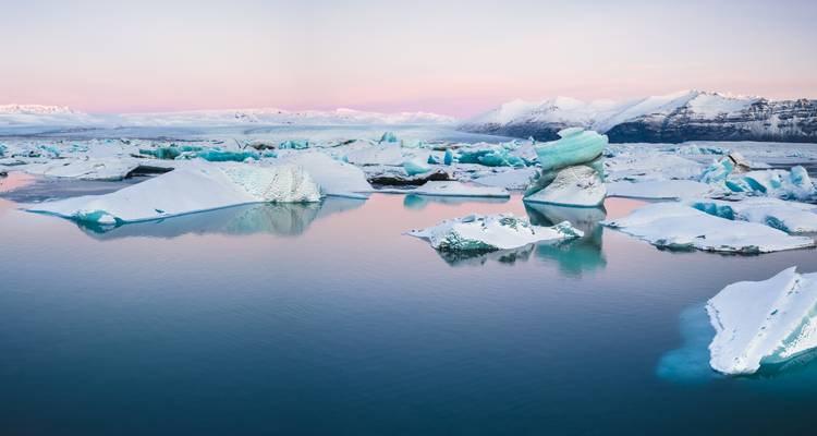 Icy blue lagoon scattered with drifting icebergs beneath pastel sunrise skies.