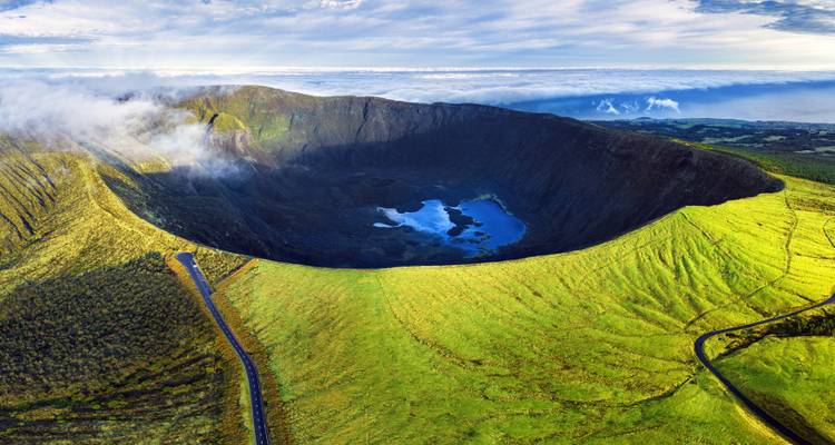 Aerial panorama of a vast green volcanic caldera with a blue lake nestled inside and clouds spilling over the rim.