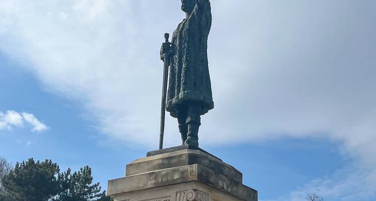 Bronzen standbeeld van een historische figuur gemonteerd op een voetstuk onder een gedeeltelijk bewolkte hemel.