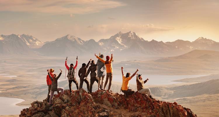 Eine jubelnde Gruppe hebt siegreich ihre Arme auf einem felsigen Gipfel, während sanftes Morgenlicht entfernte Bergrücken und Seen beleuchtet.