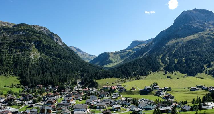 Ein malerisches Alpendorf schmiegt sich in ein grünes Tal zwischen steilen bewaldeten Bergen unter einem klaren Himmel.