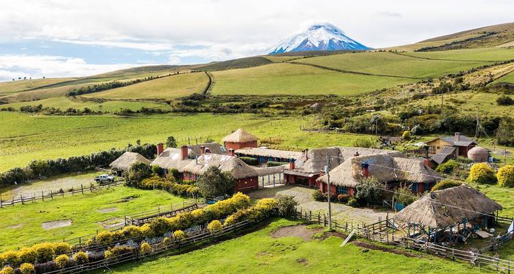 Luftaufnahme einer traditionellen Anden-Hacienda mit Strohdächern inmitten sanft geschwungener grüner Weiden, mit dem schneebedeckten Vulkan Cotopaxi im Hintergrund.