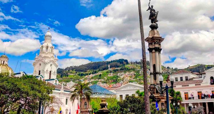 Farbenfrohe Aussicht auf Quitos historisches Zentrum mit weißen Kolonialkirchen, Denkmälern and grünen Hügeln unter einem strahlend blauen Himmel mit dicken Wolken.