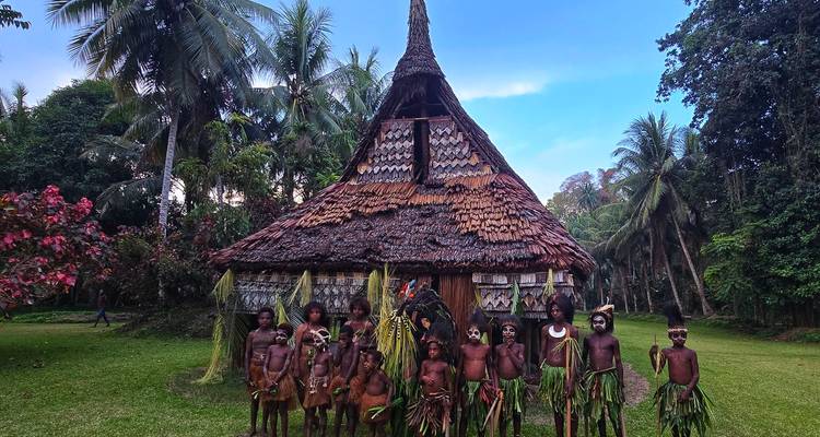 Rij traditionele Sepik-dorpelingen in bladrokjes poseren voor een huis met rieten dak en steile kap.