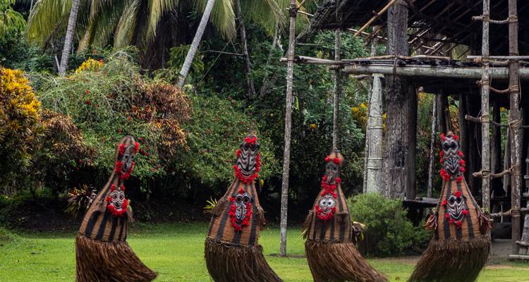 Gesneden houten krokodil geestfiguren met rode maskers staan op een grasrijke open plek tussen palmbomen.
