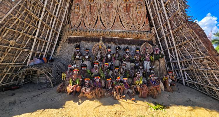 Grote groep van uitgebreid beschilderde stamleden zit onder een torenhoge geweven geestenhuis gevel.