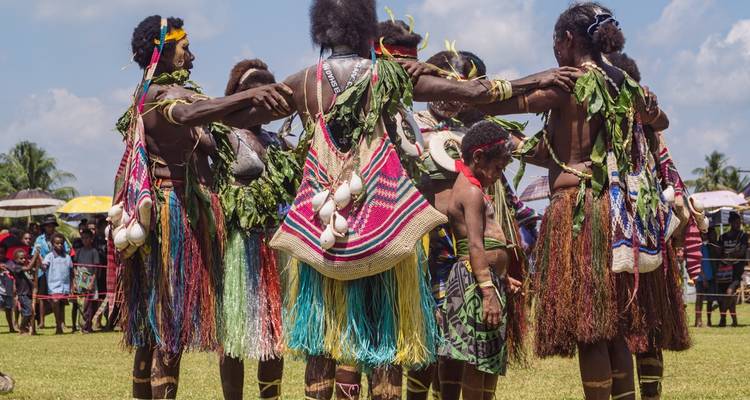 Dansers haken hun armen in elkaar in een cirkel en dragen kleurrijke rokken, bladeren en schelpornamenten tijdens een veldfestival.