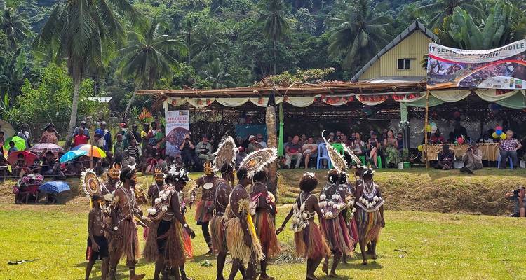 Traditionele gekostumeerde artiesten verzamelen zich op het gras terwijl toeschouwers kijken vanaf schaduwrijke tribunes bij een landelijke show.
