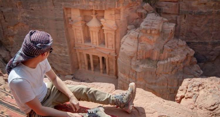 Reiziger zit op kliftrand met uitzicht op Petra's iconische Schatkamer uitgehouwen in rozerode rots