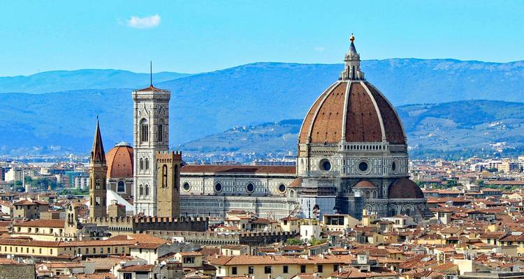 Le dôme iconique aux tuiles rouges et la façade en marbre d'une cathédrale majestueuse dominent un paysage urbain historique encadré par les montagnes.