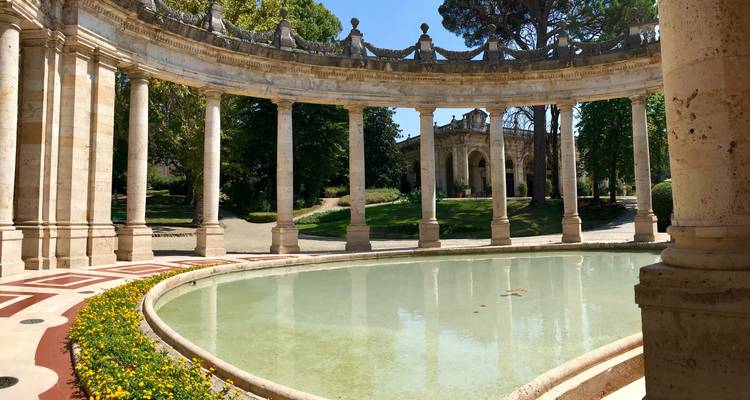 Des colonnades de pierre élégantes entourent un bassin circulaire peu profond orné de fleurs jaunes en fleur dans un parc verdoyant.